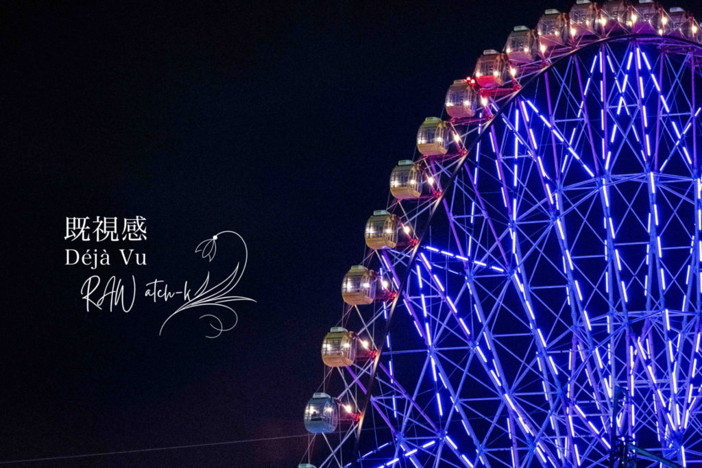 夜の暗い空を背景に、青と紫の光で照らされた観覧車の半分だけが写っている写真。止まっているように見えながら、ゆっくり回転している観覧車が、既視感と時間のずれを感じさせる。