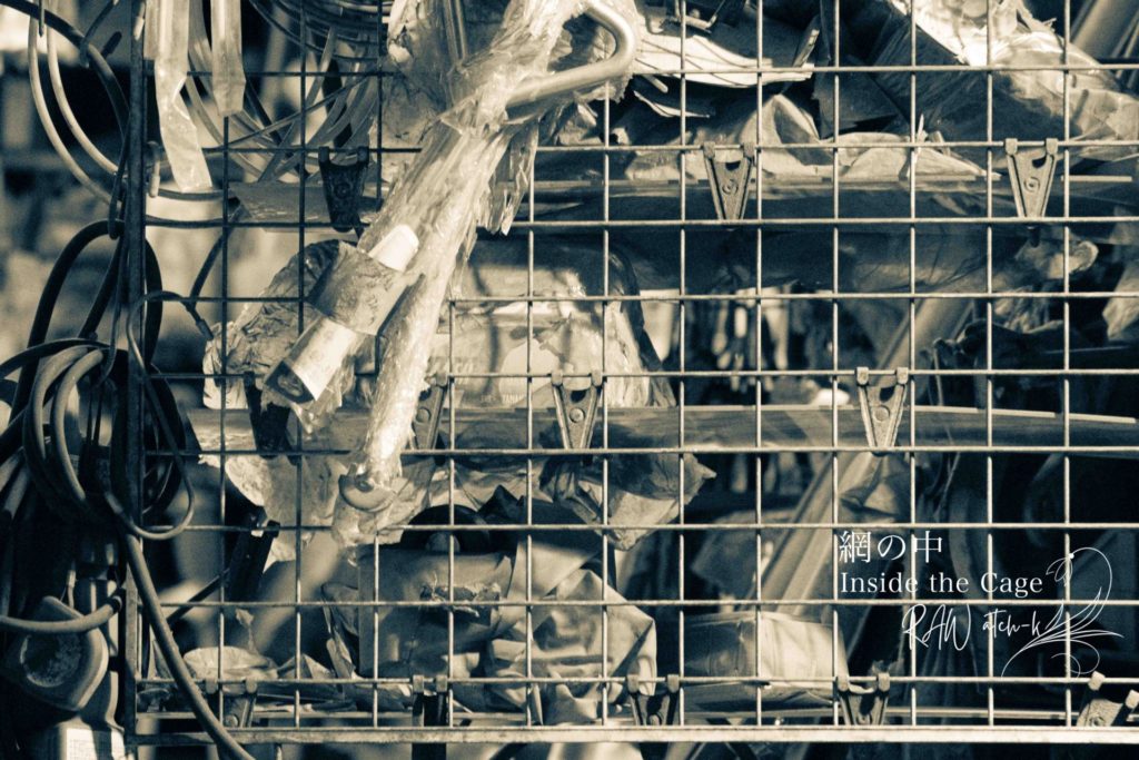 金属の網棚の中に、ビニールに包まれた配線や部品が無造作に収められ、使われるのか廃棄されるのか分からないまま静かに置かれている。